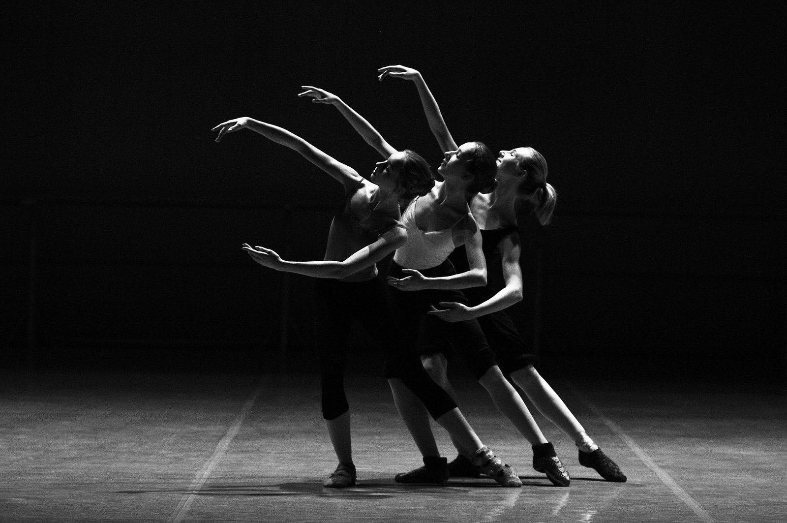 Trois danseuses en mouvement, bras levés, sur une scène plongée dans l'obscurité